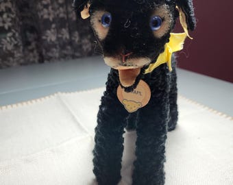 Puede incluir: Un juguete de perro de peluche negro vintage con ojos azules, una cinta amarilla y una etiqueta. El perro tiene una textura de pelaje rizado y está de pie sobre una superficie blanca. La etiqueta tiene la palabra "RAPL".