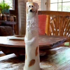 May include: A tall, cylindrical ceramic vase in an off-white finish, featuring a sculpted, three-dimensional dog figure on the front. The dog has brown ears and patches, with a textured white chest. The vase body also displays scattered brown spots.