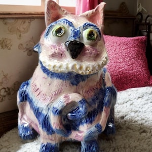 May include: A whimsical ceramic cat figurine with a striped pattern in shades of blue and pink. The cat has large green eyes, a black nose, and a white collar. The figurine is a decorative item.
