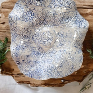 Puede incluir: Un plato de cerámica para servir con una forma orgánica única. El plato presenta un patrón de remolinos blancos y azules. El borde tiene un borde irregular. El plato está colocado sobre un trozo de madera.