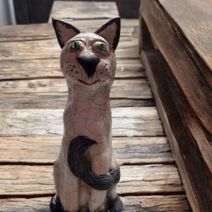 Może przedstawiać: Ceramiczna figurka kota z jasnobeżowym wykończeniem typu craquelé, z ciemnobrązowymi uszami, czarnym nosem, czarnymi łapami i czarnym ogonem owiniętym wokół przodu. Kot ma jasne zielone oczy i mały, namalowany na czerwono pyszczek.