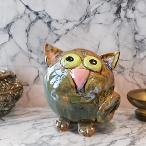 Może przedstawiać: Okrągła, glazurowana ceramiczna figurka kota z nakrapianym wykończeniem w kolorze oliwkowym i brązowym. Kot ma dwoje dużych, wyraźnych jasnozielonych oczu z czarnymi źrenicami, trójkątny różowy nos, małe spiczaste uszy oraz zakręcony ogon.