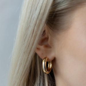 May include: Gold hoop earrings with a thick, rounded design. The earrings are worn in the ear of a person with blonde hair.