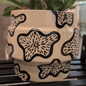 May include: Cream ceramic plant pot with a black floral design, containing a green plant and pink flowers. The pot has a distinctive shape and is placed on a black metal shelf. A decorative item for the home.