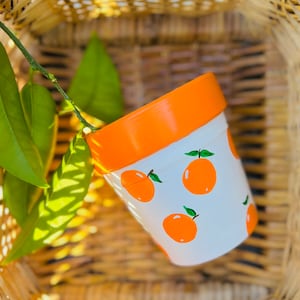 Puede incluir: Una maceta de cerámica blanca con un borde naranja y un patrón de rodajas de naranja pintadas en la parte delantera. La maceta está sentada en una cesta tejida.