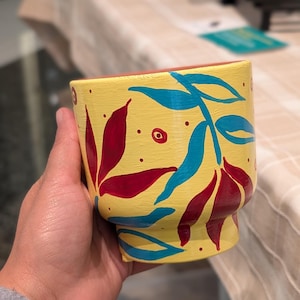 May include: A hand-painted yellow ceramic planter with a red interior. The planter is decorated with red and blue leaf designs. The planter has a wide, slightly flared shape and a small base.
