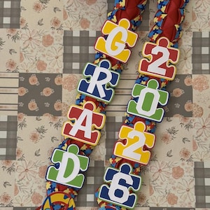 Könnte beinhalten: Ein Abschluss-Lei mit rotem geflochtenem Band und Puzzleteil-Akzenten. Das Lei schreibt "GRAD 2026" mit farbenfrohen Puzzleteil-Buchstaben und -Zahlen. Das Lei hat auch Bandakzente in Form von Puzzleteilen.