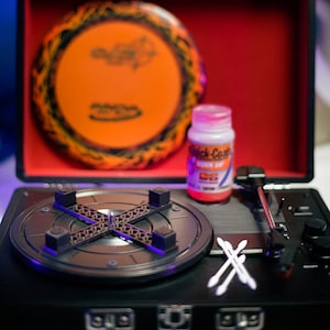May include: A black record player with a turntable, tonearm, and control knobs. A bright orange disc with black flames is on top of the record player. A small bottle of clear liquid with a red label that says "Quick-Coat" is next to the turntable. Two white cotton swabs are on the right side of the turntable.