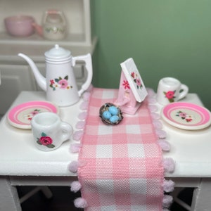 May include: A pink and white gingham table runner with white pom-poms along the edges. The runner is on a white table with a white pitcher, two white mugs with pink floral designs, two plates with pink floral designs, a small birdhouse, and a nest with blue eggs.