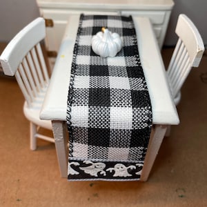 May include: Miniature white table with two matching chairs, a black and white checkered table runner, and a white pumpkin. The table runner has a decorative border with ghost silhouettes. The scene evokes a Halloween or autumn theme.