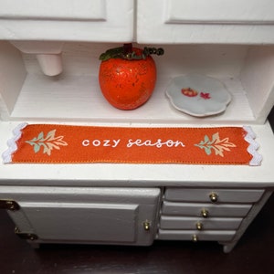 May include: A white miniature kitchen cabinet with three drawers and a shelf. The shelf has a small orange pumpkin, a white plate with a small orange pumpkin, and a decorative orange runner with the text "cozy season".