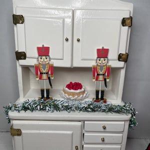 May include: White wooden hutch with two doors and four drawers. The hutch is decorated with a garland of green and white tinsel, two nutcracker figurines, and a cake with red berries on top.