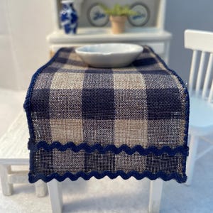 May include: Miniature dollhouse table setting with a navy blue and tan plaid table runner. A white ceramic bowl sits on the runner. The runner has a navy blue scalloped edge. A white chair and cabinet are in the background.