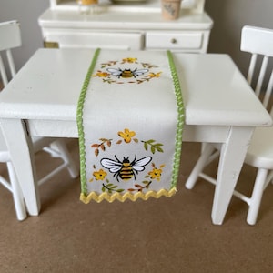 May include: A white table runner with a green border and yellow stitching. The runner features a bee and floral design.