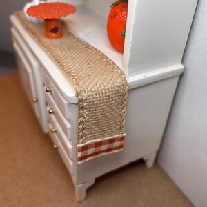 May include: Miniature white cabinet with drawers, adorned with a woven burlap runner. An orange pumpkin and a small orange table with a purple base sit on top. The runner has a red and white checkered border.