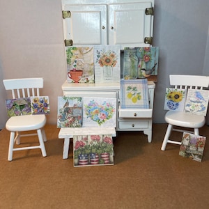 May include: Miniature white cabinet with several small framed art prints. The prints feature floral and nature scenes, including sunflowers, birds, and lemons. Two small white chairs flank the cabinet, each holding additional art prints. The overall theme is a charming, decorative display.