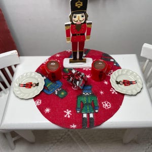 May include: A miniature Christmas table setting with a red tablecloth, two white plates with nutcracker designs, two red mugs, a wrapped gift, and a nutcracker figurine.
