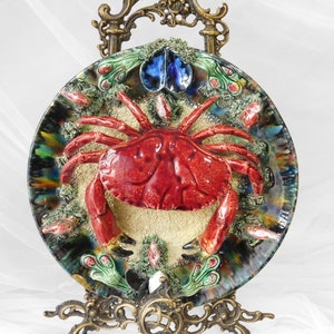 Può includere: Un piatto in ceramica con un granchio rosso in rilievo su uno sfondo sabbioso. Il piatto è decorato con alghe verdi, conchiglie blu e bianche e una glassa iridescente colorata.