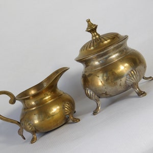 Peut inclure: Deux pièces de service antiques en laiton. L'une est une crémière avec une anse et un bec courbés, l'autre est un sucrier avec couvercle. Les deux pièces ont un aspect vieilli et terni et sont soutenues par trois petits pieds.