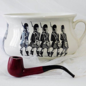 May include: A white ceramic chamber pot with a black and white illustration of a line of men in kilts. A red and black pipe is in the foreground.