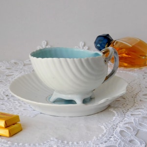 Puede incluir: Una taza de té y un platillo blancos con un interior azul y un diseño con forma de concha. La taza tiene un borde dorado y se encuentra sobre un mantel de encaje blanco.