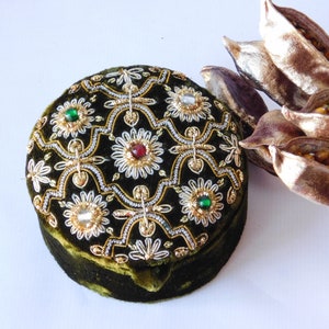May include: A round, green velvet box with gold and silver embroidery and colourful jewels. The box has a decorative pattern of flowers and geometric shapes.
