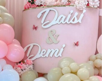 May include: A pink backdrop with the names "Daisi & Demi" in white script, adorned with butterflies and floral arrangements. Balloons in shades of pink, white, and beige surround the backdrop, creating a festive atmosphere.