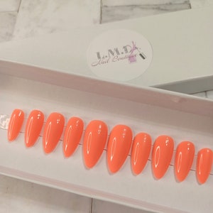 May include: A set of ten peach-colored almond-shaped press-on nails in a white box. The box has a sticker with the text "I.M.D. Nail Boutique".