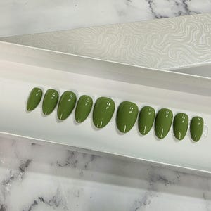May include: A set of ten olive green press-on nails with a glossy finish and an almond shape, arranged in a row inside a white rectangular display box. The nails vary in size from largest to smallest, showcasing a smooth, reflective surface.