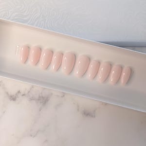 May include: A set of light pink, oval-shaped press-on nails arranged in a white box. The nails have a glossy finish and are displayed against a white and marble background. The box has a textured, wave-like pattern.