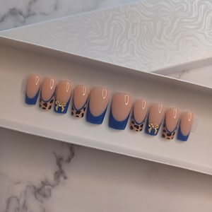 May include: A set of ten artificial nails in a white box. The nails have a nude base with a blue French tip and leopard print accents. Some nails feature a gold bow. The nails are designed for cosmetic use.