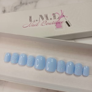 May include: A set of ten light blue press-on nails with glitter accents. The nails are arranged in a row in a white box with the text "L.M.D. Nail Boutique" on the lid.