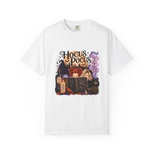 Może przedstawiać: Biały t-shirt z krótkim rękawem z nadrukiem graficznym przedstawiającym trzy postacie za księgą zaklęć z okiem na okładce. Tekst głosi Hocus Pocus, It's just a bunch of Hocus Pocus oraz Better Witches Together. Zawiera butelkę z miksturą i miotłę.