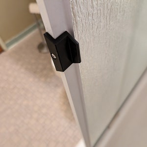 May include: Close-up of a black shower door handle with a silver screw. The handle is attached to a white shower door frame.