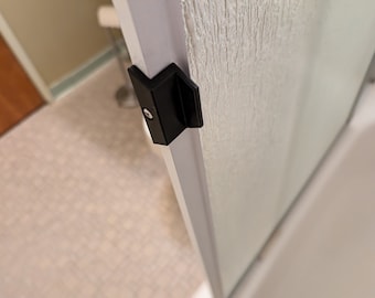 May include: Close-up of a black shower door handle with a silver screw. The handle is attached to a white shower door frame.