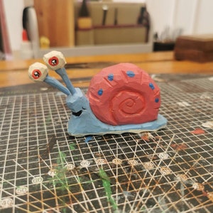May include: A wooden snail ornament, hand-carved with a blue body and a red shell adorned with blue dots. The snail has two eye stalks with googly eyes and a smiling face. It rests on a grid-patterned surface.
