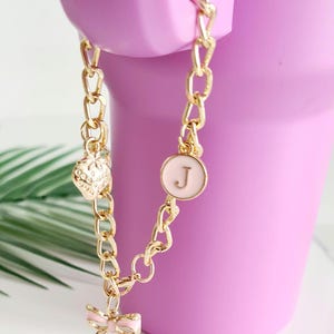 May include: A gold chain charm bracelet with a pink bow, a pink heart with a floral design, and a pink circle with the letter "J" in gold.
