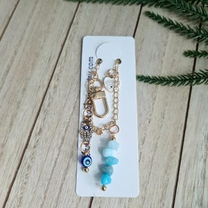 May include: A gold-toned metal keyring featuring a lobster clasp, a hamsa hand charm with clear rhinestones, a small white heart charm with an eye graphic, a dark blue evil eye bead, and a strand of light blue and turquoise stone beads.