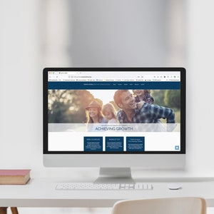 May include: A computer screen showing a website with the words "ACHIEVING GROWTH" and a family image. The computer sits on a white desk, with a small, aged book beside it. The website's header is dark blue.