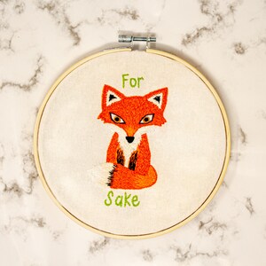 May include: Embroidered hoop art featuring a fox with the text "For Sake" in green thread.