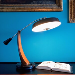 Könnte beinhalten: Eine Retro-Schreibtischlampe mit geschwungenem Holzfuß und schwarzem Lampenschirm. Die Lampe steht auf einer schwarzen Oberfläche neben einem offenen Buch. Der Hintergrund ist eine türkisfarbene Wand mit gerahmten Kunstwerken.
