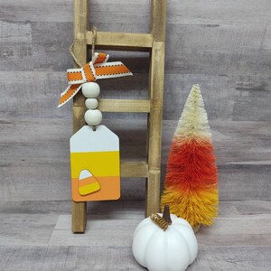 May include: A rustic wooden ladder with a candy corn tag, a white pumpkin, and a candy corn-colored tree. The tag has a white, yellow, and orange design. The bow is orange and white. The tree is orange, yellow, and white.
