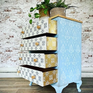 May include: A light blue chest of drawers with four drawers. The drawers are decorated with a white and gold geometric pattern. The chest of drawers is sitting on a wooden floor.