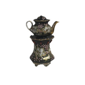 Puede incluir: Una tetera de porcelana negra con un diseño floral dorado y blanco se asienta sobre un soporte a juego con tres patas. La tetera tiene una tapa y un pico.