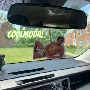 Op de afbeelding: Een autoluchtverfrisser met een foto van een man in een witte handdoek. De luchtverfrisser hangt aan de achteruitkijkspiegel. De tekst "COOLMODA!" is zichtbaar in de afbeelding.
