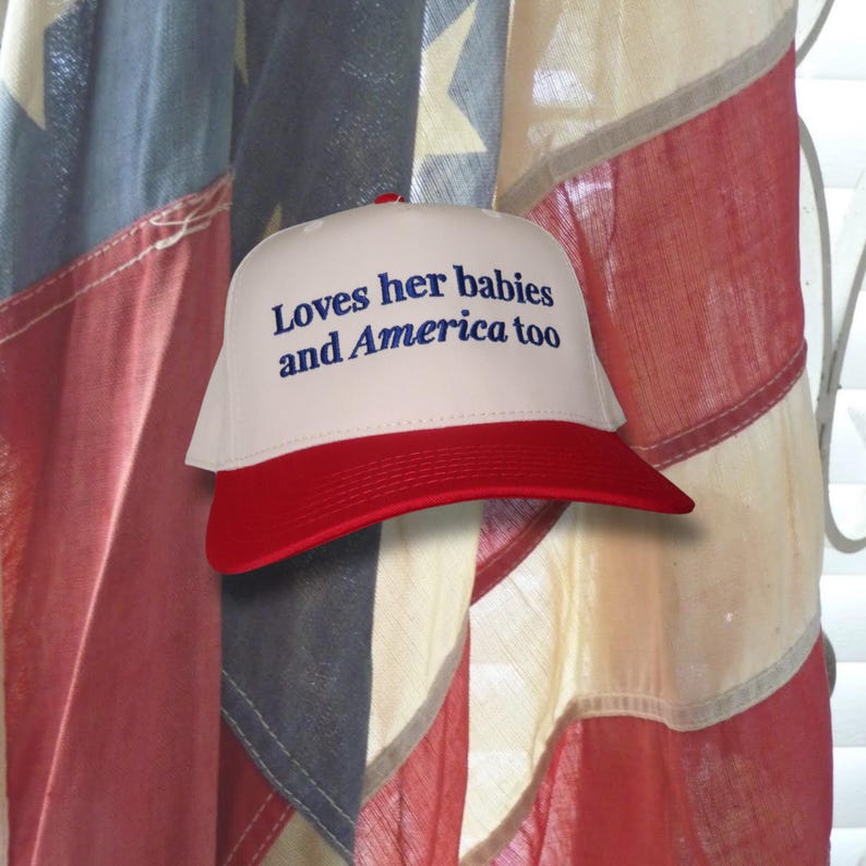 Puede incluir: Gorra de b&eacute;isbol blanca y roja con el texto "Loves her babies and America too" en azul. La gorra tiene una visera roja y un bot&oacute;n rojo en la parte superior. El fondo presenta una bandera americana.