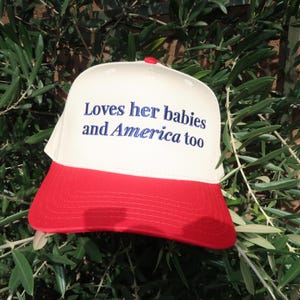 embroidered canvas cream and red hat in royal blue thread that says Loves her babies and America too