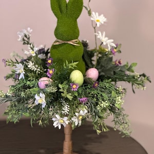 May include: A green moss bunny sits in a centerpiece of white and purple flowers, green foliage, and pastel Easter eggs on a wooden pedestal.