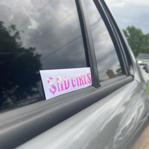 Puede incluir: Una pegatina rectangular con el texto "$AD GIRLS" en una fuente pixelada. El texto está en tonos de rosa y morado con un símbolo de dólar. La pegatina está adherida a la ventana de un coche plateado.