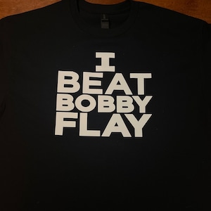 May include: Black t-shirt with the white text "I BEAT BOBBY FLAY" in a bold, blocky font. The shirt is made of a soft material and is laid flat on a wooden surface. The collar and shoulder seams are visible.